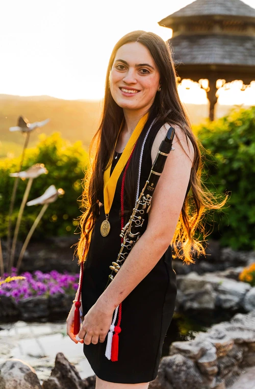 The picture depicts a girl standing outside with the sun glowing behind her. She is wearing a black dress and has graduation cords and a medal around her neck. She has a clarinet in her arm. 